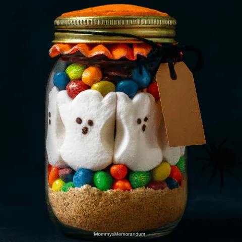 Halloween S&rsquo;mores in a Jar with ghost Peeps, M&Ms, and graham cracker crumbs layered in a mason jar with orange fabric lid and spooky tag.