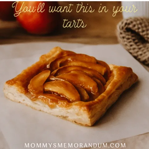 A golden caramel apple tart sits on parchment paper atop a rustic wooden table. The tart features flaky puff pastry layered with caramel-glazed apple slices arranged in neat rows. In the background, red apples, a cozy knit blanket, and a ceramic mug evoke a warm fall aesthetic. Text overlay reads: &ldquo;The Caramel Apple Tart &mdash; You&rsquo;ll want this in your tarts.