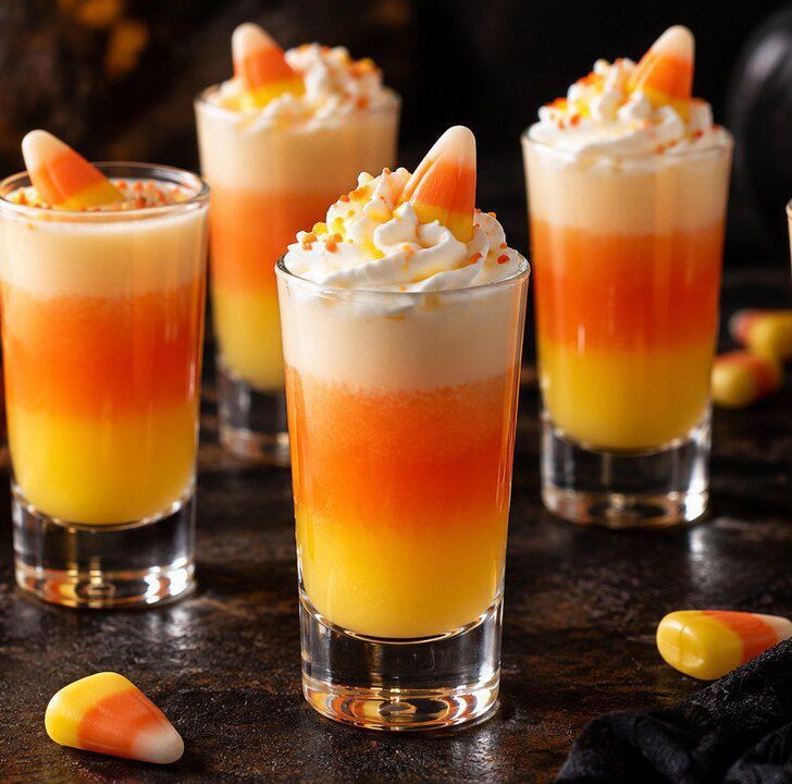 Five Candy Corn Shooters in clear shot glasses with orange, yellow, and cream layers, topped with whipped cream and candy corn for Halloween.