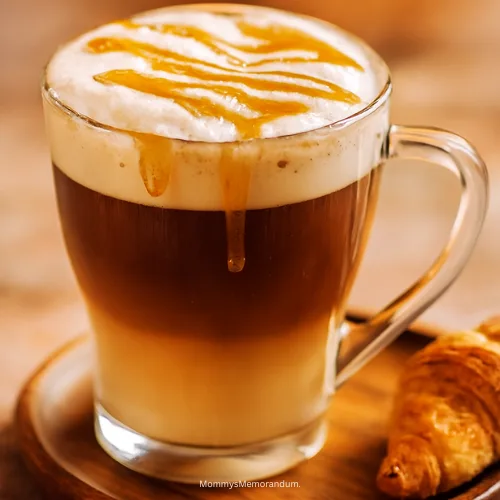 A cozy caramel macchiato served in a clear glass mug with visible espresso and milk layers, topped with frothy foam and caramel drizzle. A croissant sits beside it on a rustic wooden tray, bathed in warm morning light.