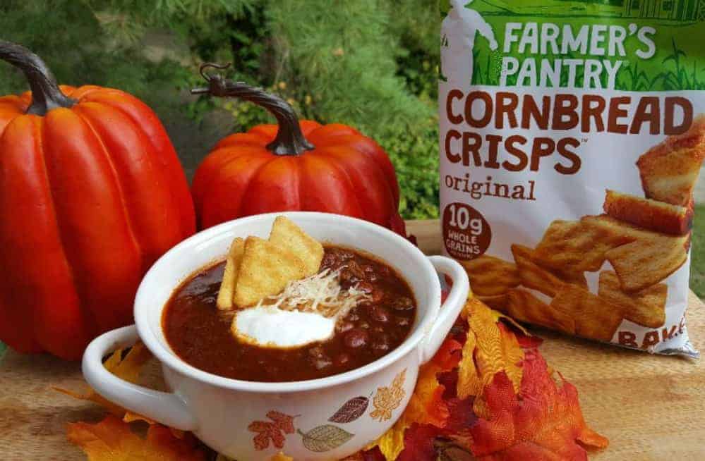 Bowl of Coca Cola Chili topped with sour cream and cornbread crisps, surrounded by decorative pumpkins and a bag of Farmer's Pantry Cornbread Crisps.