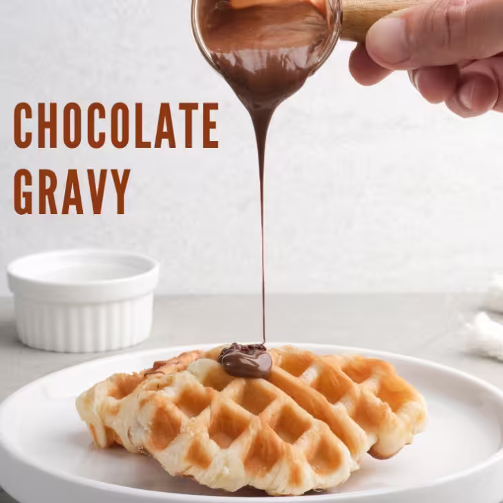 A close-up of a hand pouring chocolate gravy over a fresh waffle on a white plate.