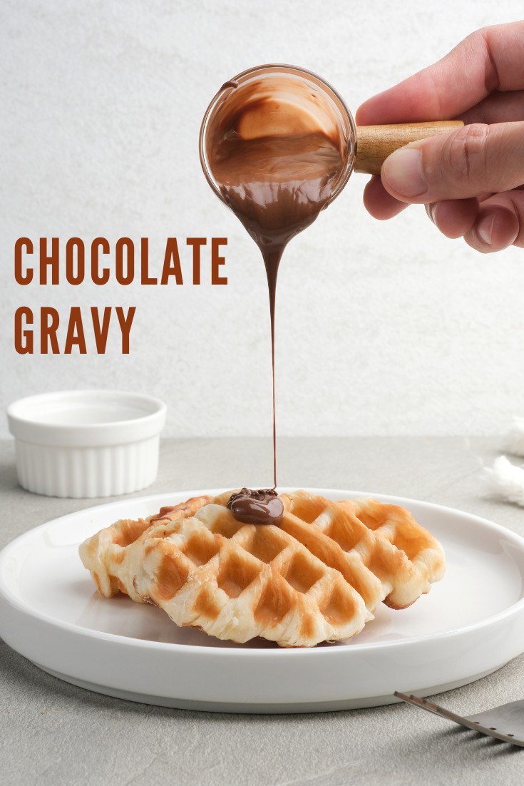 A close-up of a hand pouring chocolate gravy over a fresh waffle on a white plate.