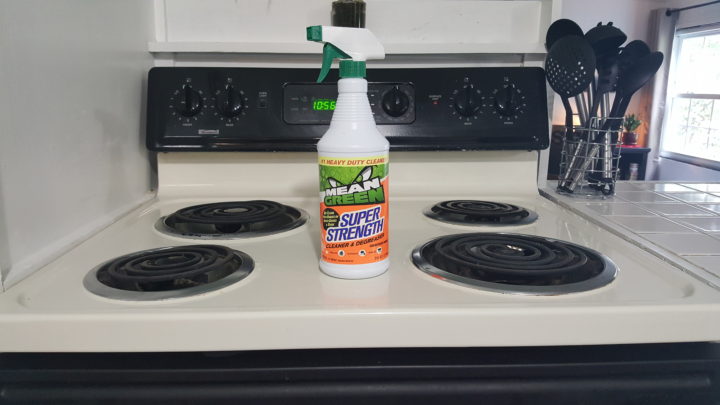A bottle of Mean Green Super Strength Cleaner stands on a white kitchen stove, near utensils and a tiled backsplash.