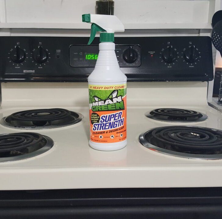 A bottle of Mean Green Super Strength Cleaner stands on a white kitchen stove, near utensils and a tiled backsplash.