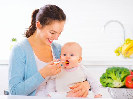 woman in blue cardigan feeding baby with broccoli resting on the table.