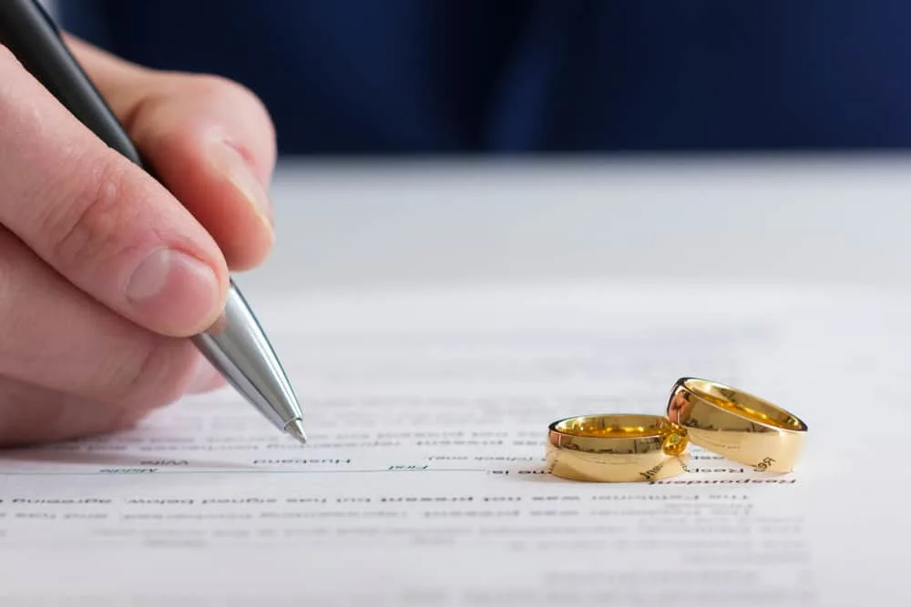 Hand signing divorce papers with two wedding rings placed on the document