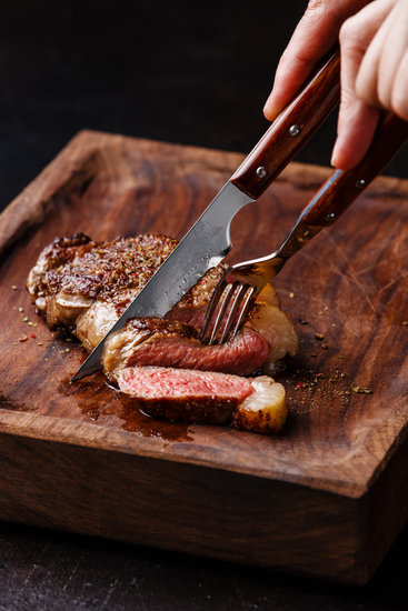 Sliced grilled meat steak New York Striploin with knife and fork in male hands on wooden board on black background close-up