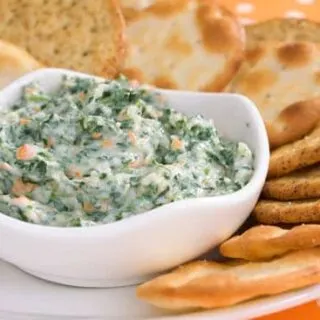 Slow cooker cheesy spinach and artichoke dip in a white bowl, served with assorted crackers on a plate