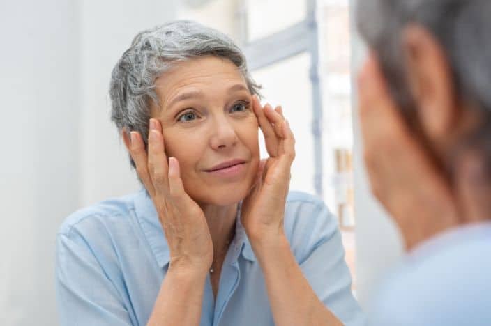 Beautiful senior woman checking her face skin and looking for blemishes. Portrait of mature woman massaging her face while checking wrinkled eyes in the mirror. Wrinkled lady with grey hair checking wrinkles around eyes, aging process. wondering at what age most health problems start