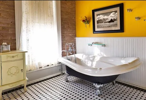 A modern contemporary classic bathroom design, furnished with a classic painted cabinet, sepia toned picture on the wall, exposed brick wall and a window, a claw foot bath tub and black and white tile pattern on the floor. Photographed in horizontal forma