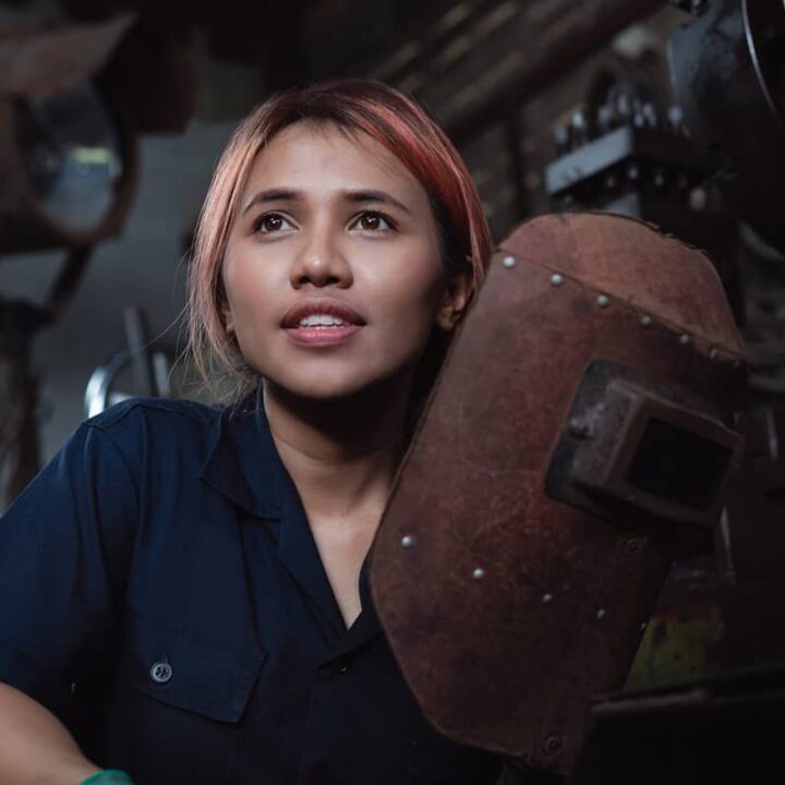 Diverse female industrial engineer holding welding helmet after work shift - Young Asian factory metal worker taking a break - Hispanic apprentice woman learning new skills on internship training