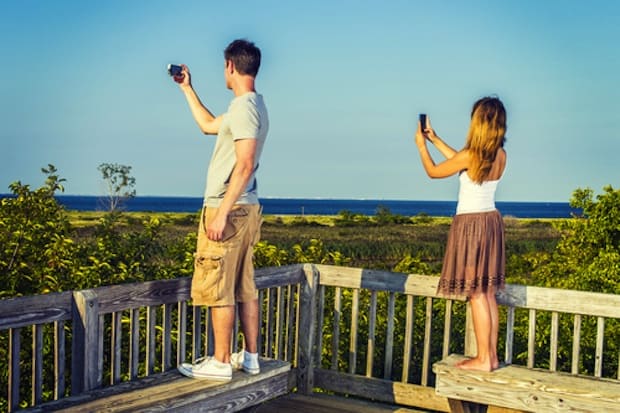 Traveling couple video recording scenery of beach on remote location, using mobile phones, guy wearing gray T shirt, yellow pants, white sneakers, girl wearing golden brown skirt, white top, barefoot.'n