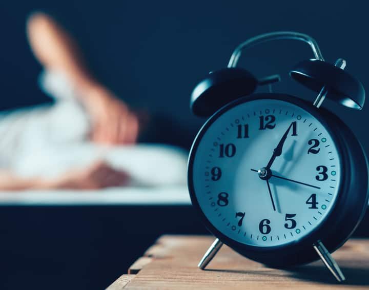 Sleeping disorder or insomnia concept, selective focus of vintage clock in bedroom and out of focus male person trying to fall asleep in bed