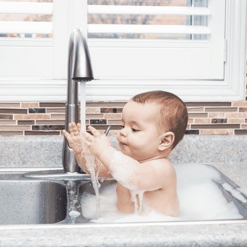 Portrait of cute Caucasian funny baby girl boy with dark black eyes sitting in big kitchen sink with water and foam near window looking away, lifestyle everyday concept