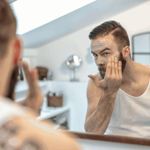 reflection in the bathroom mirror of a young bearded man applying a face mask to his skin in a skincare and hygiene concept