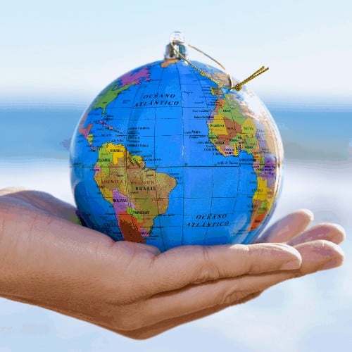 closeup of a young caucasian man with a world globe with a golden string as a christmas ball in his hand, with the ocean in the background. Depicting moving abroad.