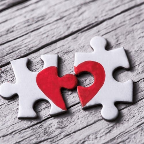 closeup of two separated pieces of a puzzle which together form a heart on a white rustic wooden surface, depicting the idea of rupture or cooperation.