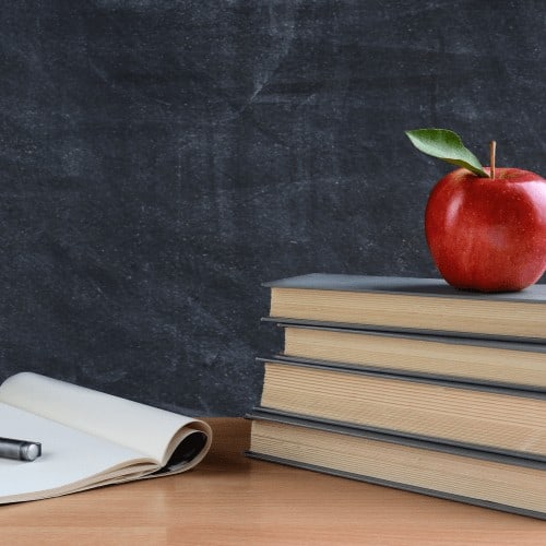 Closeup of a teachers desk with books, paper and pen and a red apple in front of a chalkboard