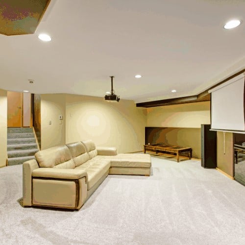 Beige basement movie room features a leather sectional under a ceiling mounted movie projector.