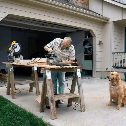 A retired craftsman adjusts a woodworking router on a temporary workbench in his suburban Missouri driveway as his Golden Retriever sits nearby.