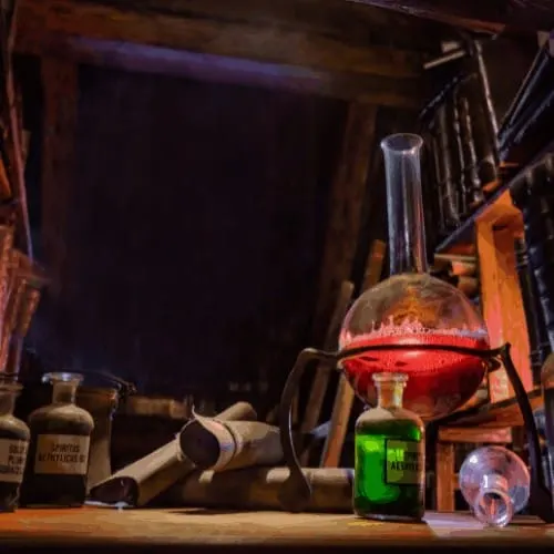 A dimly lit medieval alchemist laboratory with a red liquid in a large distillation flask, old scrolls, and various bottles.