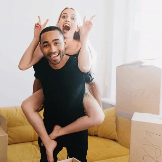 Very happy couple celebrating first day in new home, finishing packing, unpacking boxes, having fun in new apartment. Young man lifting woman on his back in room with yellow sofa.