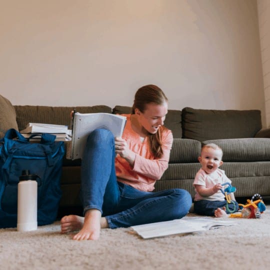 A mother and parent sits with her baby daughter on the family room floor studying for a university class. She is a hard working college student trying to earn her degree in education to improve her situation.