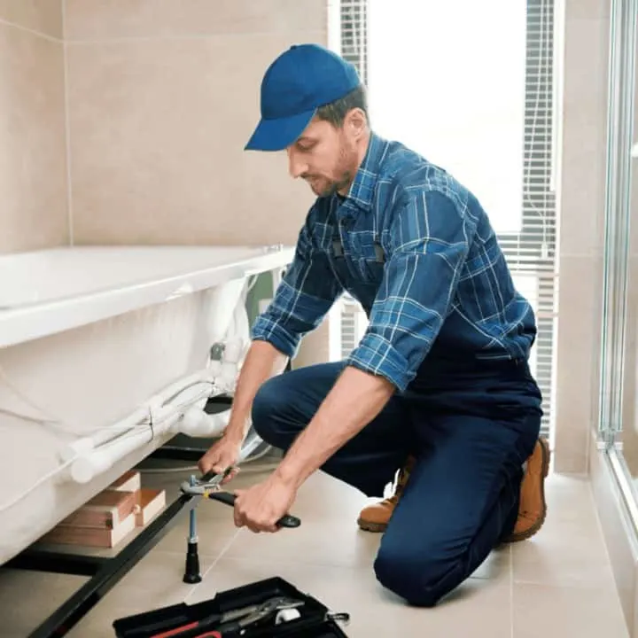 Young plumber or technician in workwear preparing detail for bathtub installation while sitting on squats