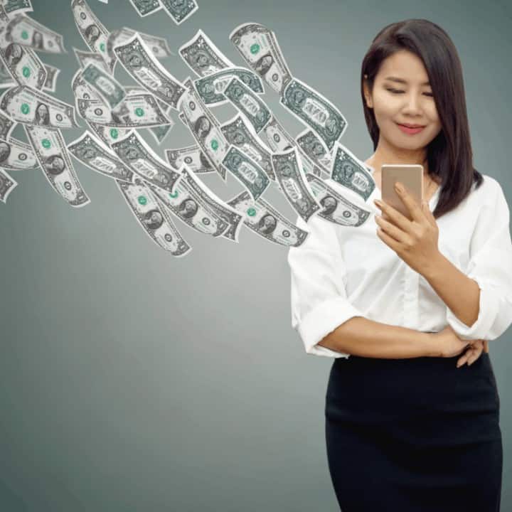 Happy Asian business woman with smart phone in hand making money online with banknotes flying in the air