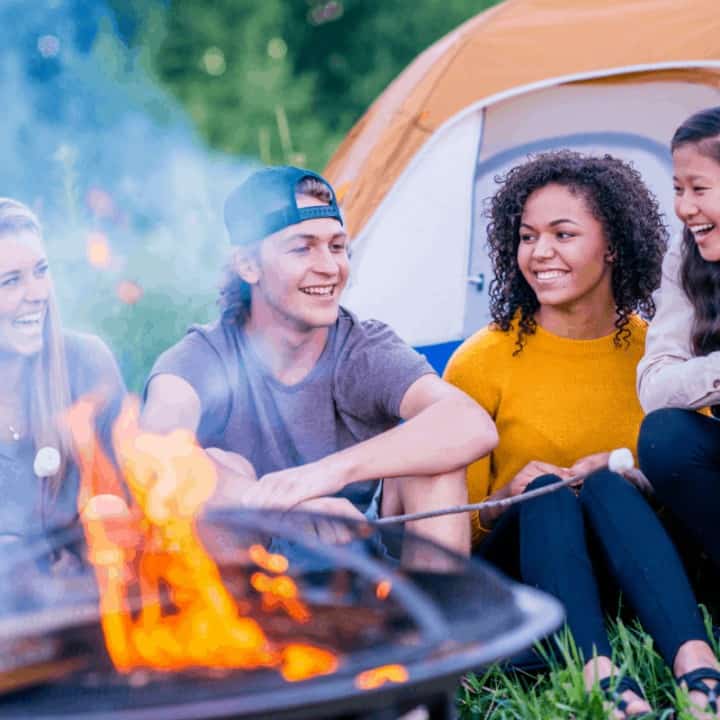 A multi-ethnic group of young adults are hanging out at a campfire. Their tent is pitched behind them. They are chatting by the fire while roasting marshmallows.