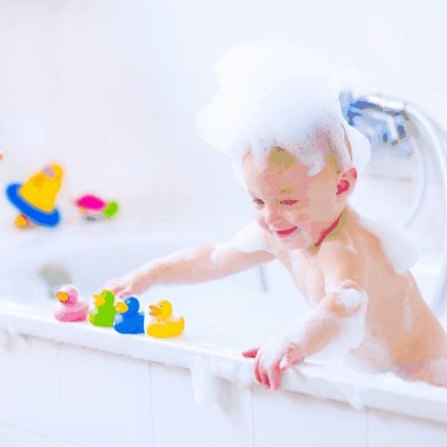 Cute little baby boy taking bath playing with foam and colorful rubber duck toys in a white sunny bathroom