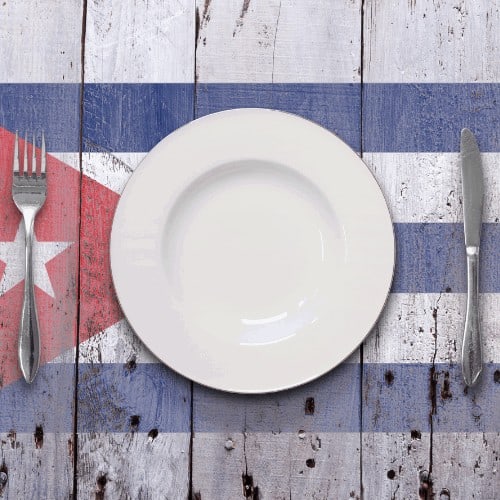 A dining scenario of a white plate and silver cutlery, shot overhead, on top of a worn rustic white wooden surface, featuring a stenciled flag.