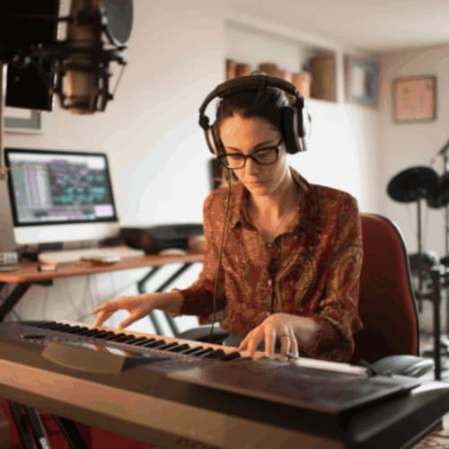 Stock photograph of a good looking, studious young woman composing music in her private home studio. She is using a computer & music keyboard to compose and is surrounded by musicians’ equipment.