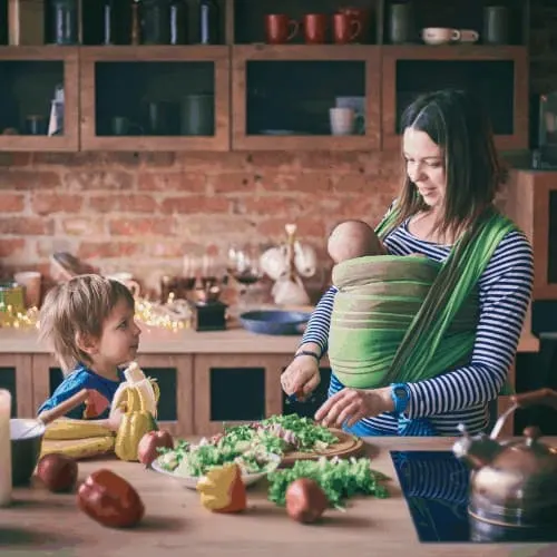 mom with newborn in momwrap on chest making a salad with toddler.