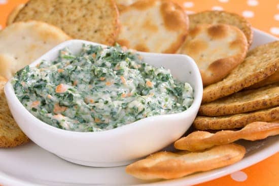 slow cooker cheesy spinach artichoke dip in white bowl on plate with dipping crackers.