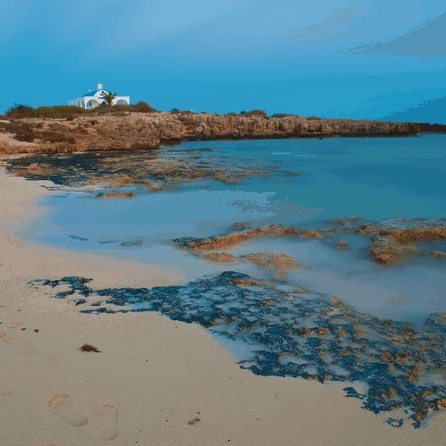 View of white Ayia Thekla chapel above sandy Poseidon Beach near ayia napa, Cyrpus. Blue sky above shallow water with reefs and footprints. Warm evening in fall. Copy space. Long exposure