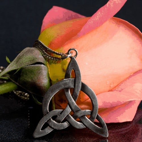 A pink and yellow rose, with a silver pendant. The design of the pendant is a celtic love knot. The surface the items are on has a black and grey texture and the background is dark. There is some reflection on the surface.