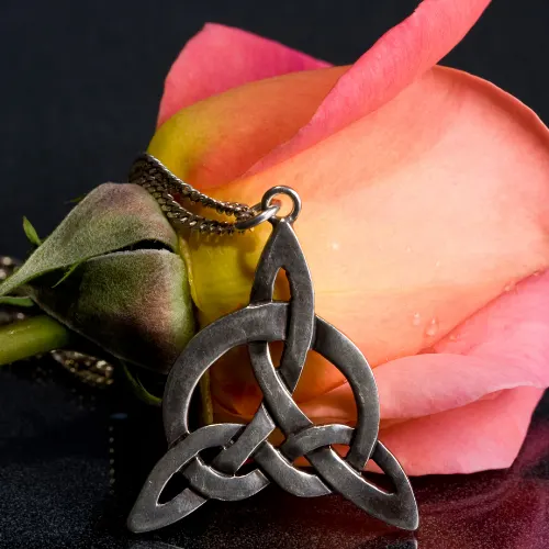 A pink and yellow rose, with a silver pendant. The design of the pendant is a celtic love knot. The surface the items are on has a black and grey texture and the background is dark. There is some reflection on the surface.