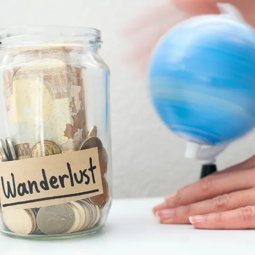 Saving up money to travel concept / Money jar with wanderlust note and rolling world map globe.