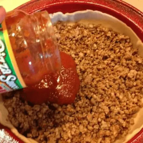 Jar of pizza sauce being poured over a layer of browned meat in a deep dish pan, assembling a copycat Pizza Hut Priazzo deep-dish pizza.
