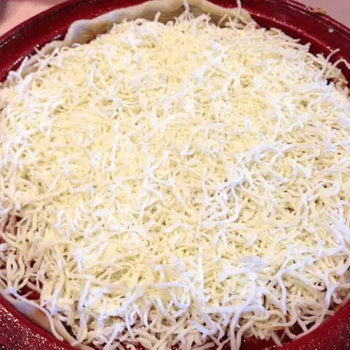 Deep dish pan filled with a final layer of shredded mozzarella cheese, completing the assembly of a copycat Pizza Hut Priazzo deep-dish pizza.