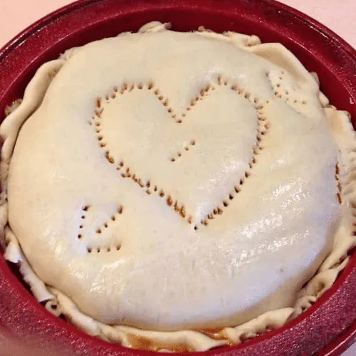 Deep dish pan with a top crust layer, featuring a heart design made with fork pricks, completing the assembly of a copycat Pizza Hut Priazzo deep-dish pizza.