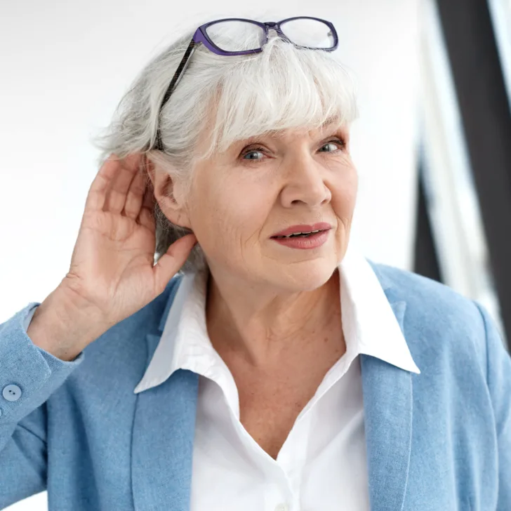 Close up elegant stylish elderly woman in formal suit having hearing problems, holding hand at her ear, trying to hear you, saying: Speak louder, please. Age, maturity, people and health concept