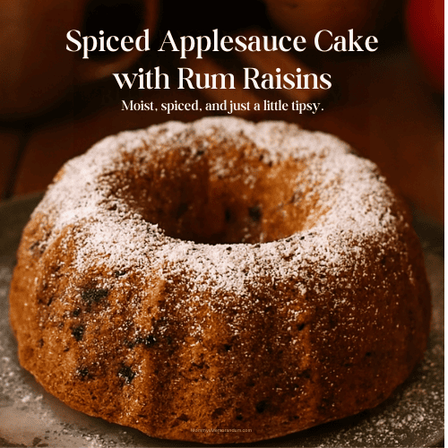 A golden-brown spiced applesauce Bundt cake dusted with powdered sugar sits on a rustic wooden table. Rum raisins are visible throughout the moist crumb. In the softly blurred background, fall elements like cinnamon sticks, apples, a pumpkin, and a steaming mug of cider create a warm, seasonal atmosphere. Overlay text reads “Spiced Applesauce Cake with Rum Raisins” and “Moist, spiced, and just a little tipsy.”