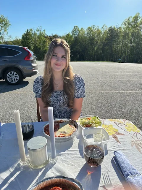 Young woman sitting at a romantic table set for a 'Me-n-U' prom proposal, featuring a beautifully laid out meal in an outdoor setting.
