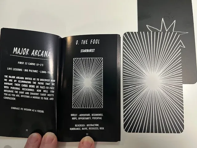 Photo of a hand holding open The Portent Tarot guidebook showing details of The Fool card with a starburst design, alongside the actual card.