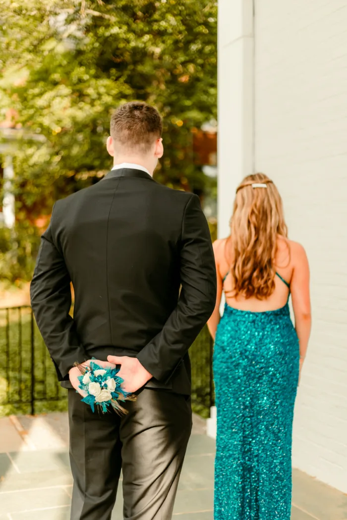 Young man in a black tuxedo holding a teal corsage behind his back for a surprise reveal to a young woman in a teal sequin dress.