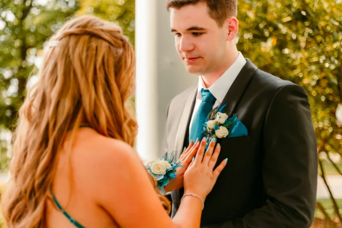 Young woman pins a teal boutonniere on her prom date's black tuxedo jacket while holding a matching teal corsage during Prom 2024.