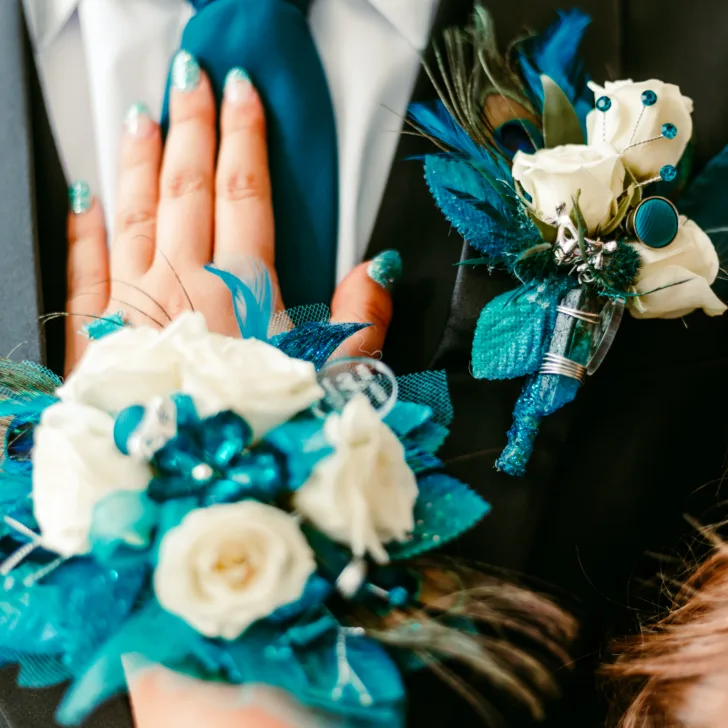 Close-up of a young woman’s hand on her prom date's chest, showing a matching teal corsage and boutonniere with peacock feathers and white roses.